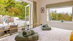 A bedroom with a bed, a window with a view of trees, a table with a vase, and a chair. - 2 Bed in Bangor (oc-66276) (Llanddeiniolen)