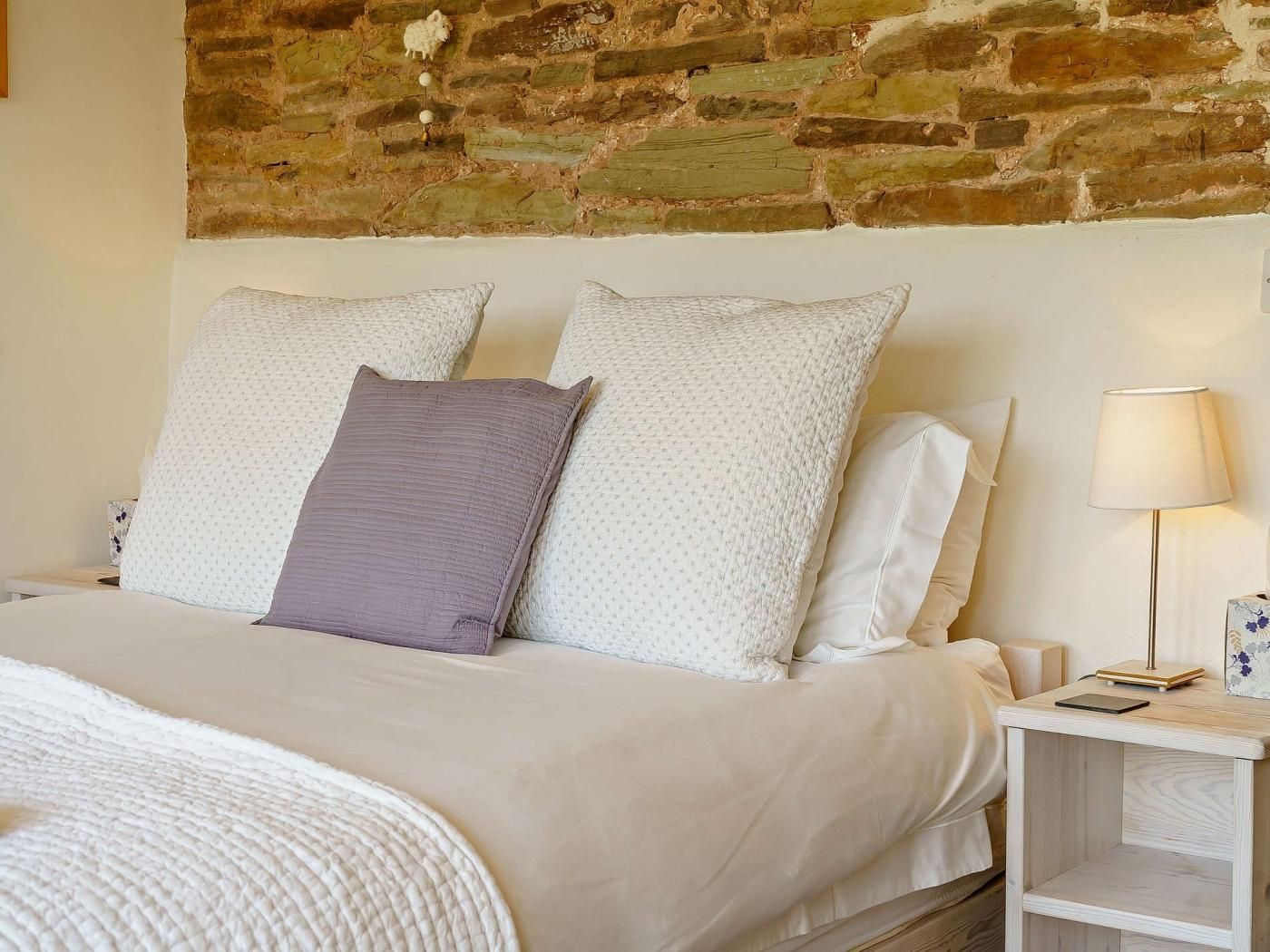 A bed with white and patterned pillows, a wooden nightstand with a lamp, and a stone wall behind.
