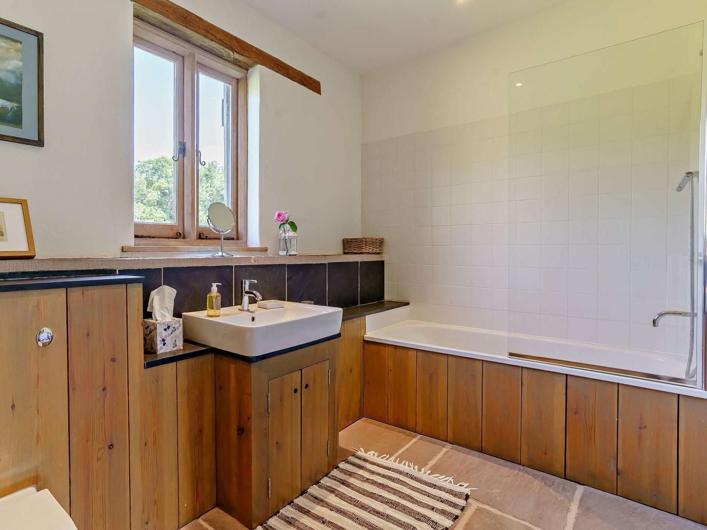 A bathroom with wooden cabinets, a white sink, a mirror, a window, and a shower area.