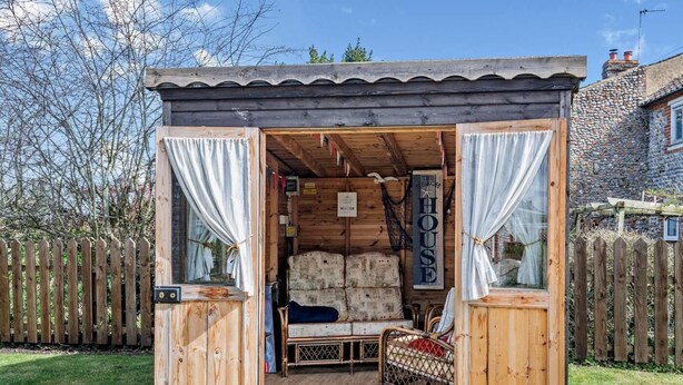 En stuga av trä med en säng, en skylt med texten ”The Beach House” och en stenmur i bakgrunden.