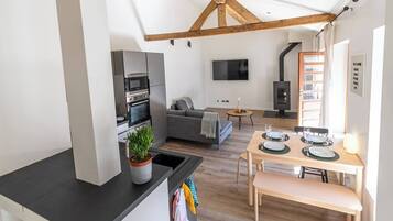 A modern kitchen with built-in appliances, a dining area with a table set for two, a grey sofa, and a TV mounted on the wall.