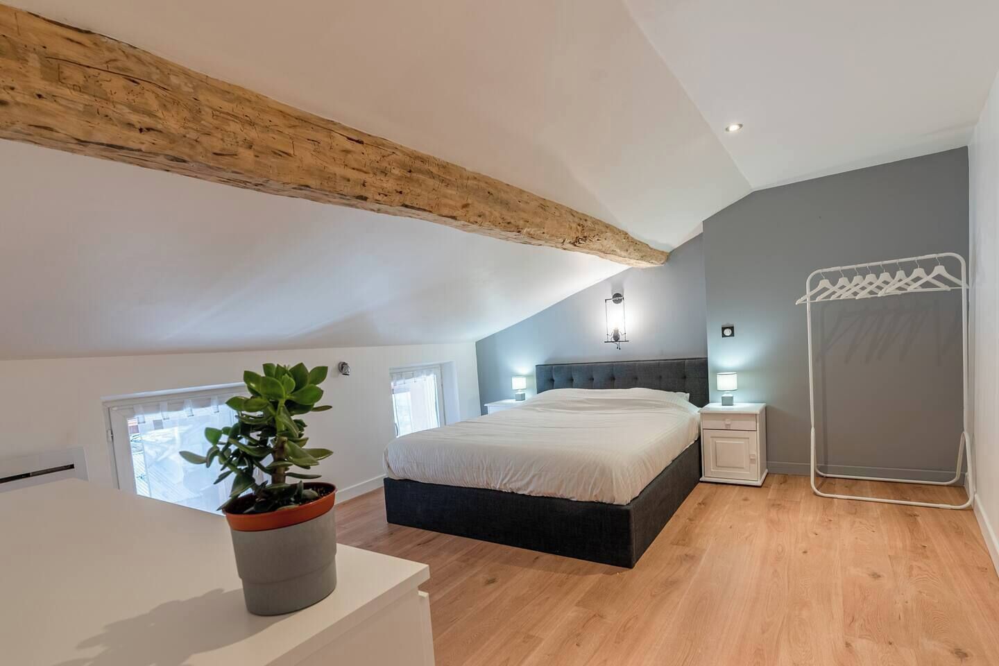 A bedroom with a sloped ceiling, a bed, bedside tables, a plant, and a hanging laundry rack.