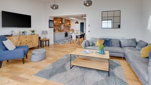 Een moderne woonkamer met een bank, salontafel en televisie. Achtergrond toont een keuken met een eethoek.