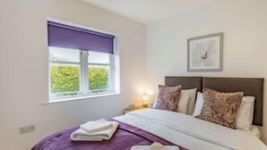 Ein Schlafzimmer mit Bett, Kissen, einem Fenster mit Blick, einem Nachttisch mit Lampe und einem gerahmten Bild an der Wand.