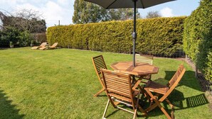 Eine Terrasse mit Tisch und Stühlen, einem Sonnenschirm und einem Hecken im Hintergrund.