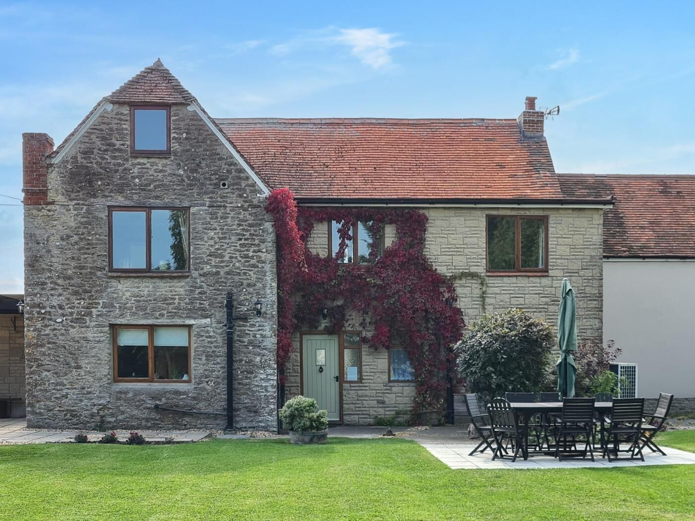 A stone house with a red-tiled roof, a green door, and a garden with a table and chairs.