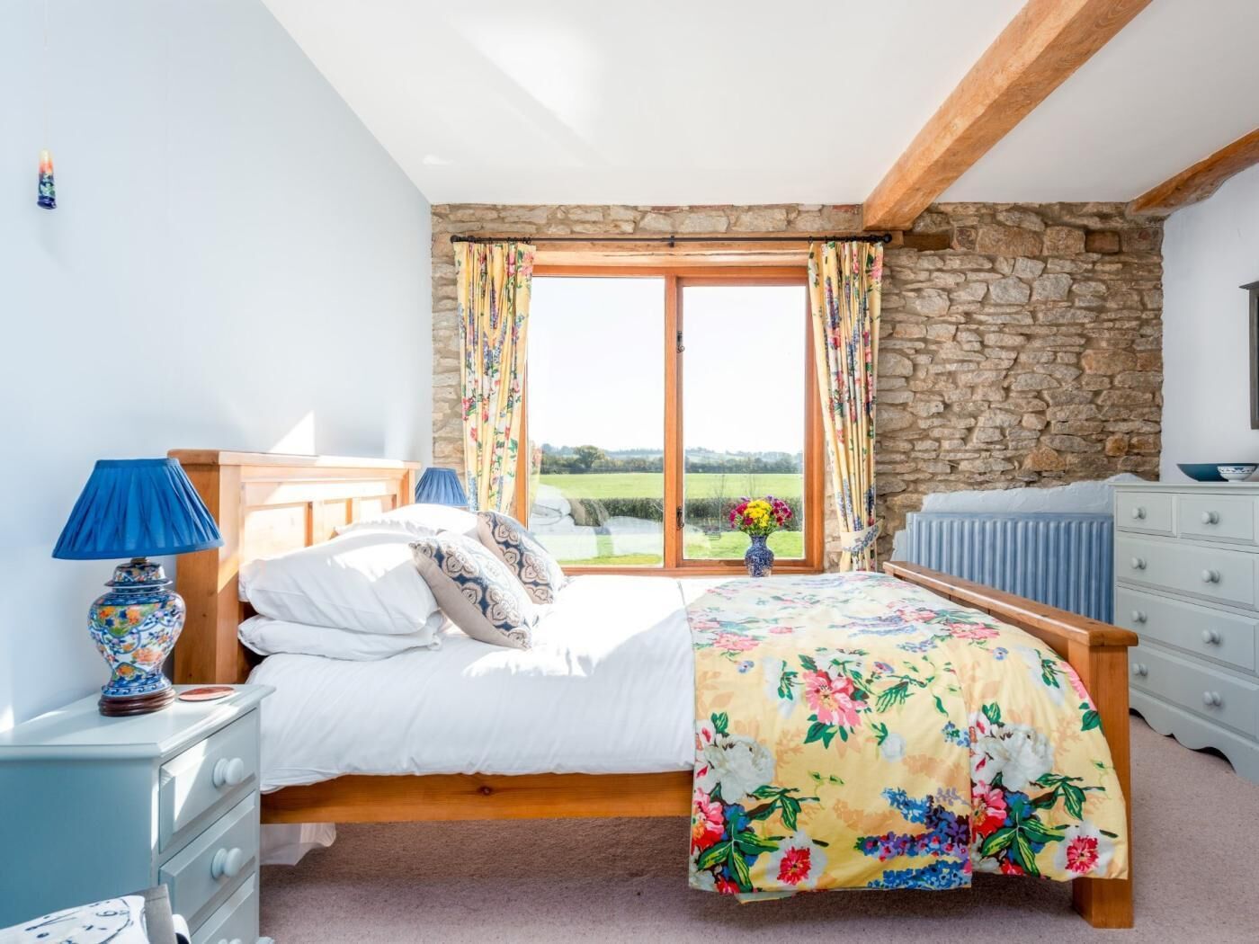 A bedroom with a bed, a dresser, a lamp, a window with curtains, a stone wall, and a view of the outdoors.