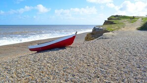 A pebble beach with a red boat, a cliff, and a few people walking. - 2 Bed in Holt (oc-ncc15) (Salthouse)