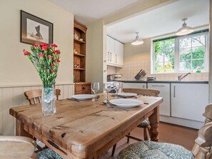 Una mesa de madera con sillas, un jarrón de flores, una cocina con armarios blancos, un ventanal y una lámpara en el techo.