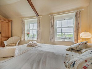 Un dormitorio con cama, dos ventanas con cortinas, una silla, una lámpara y un armario de madera.