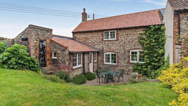 A stone house with a tiled roof, a garden with grass and flowers, and a small outdoor seating area. - 2 Bed in Holt (oc-ncc15) (Salthouse)