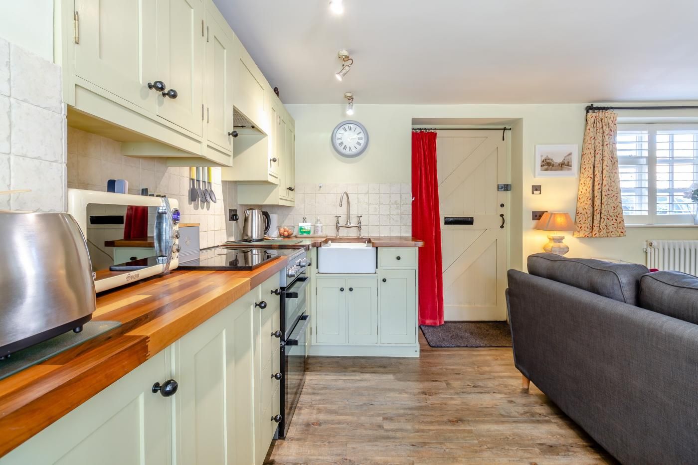 A compact kitchen with a wooden countertop, a stove, and a sink. There is a dining area with a sofa and a window with curtains.