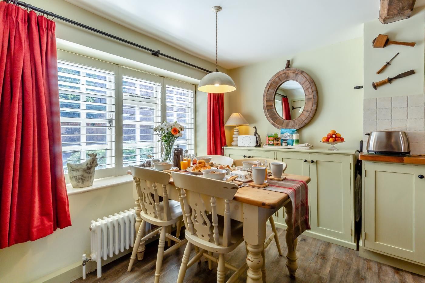A dining area with a table set for breakfast, a window with curtains, a radiator, and a kitchen area with utensils on the wall.
