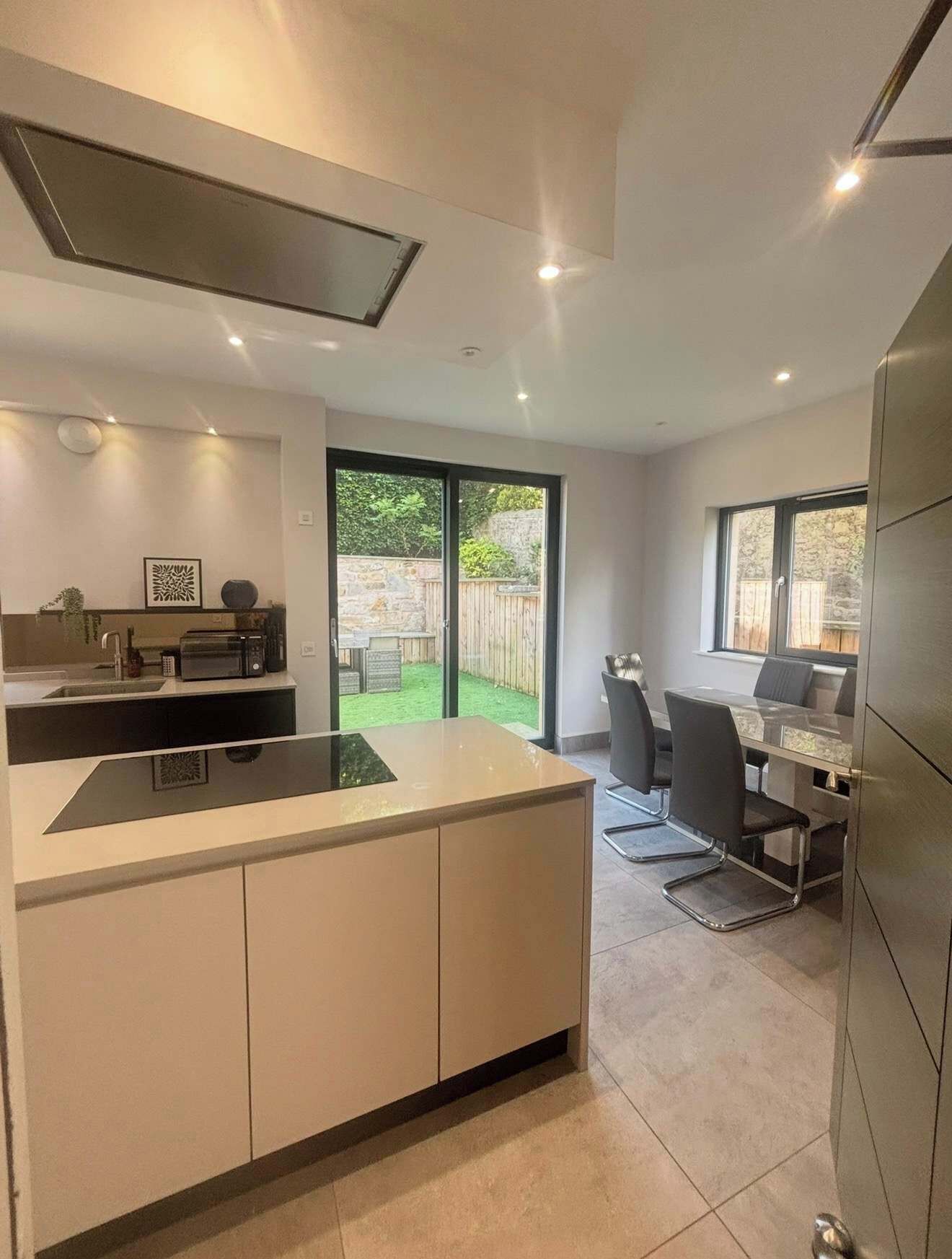 Een moderne keuken met een centraal eiland, een eethoek met een tafel en stoelen, en een uitzicht op een tuin door grote schuifdeuren.
