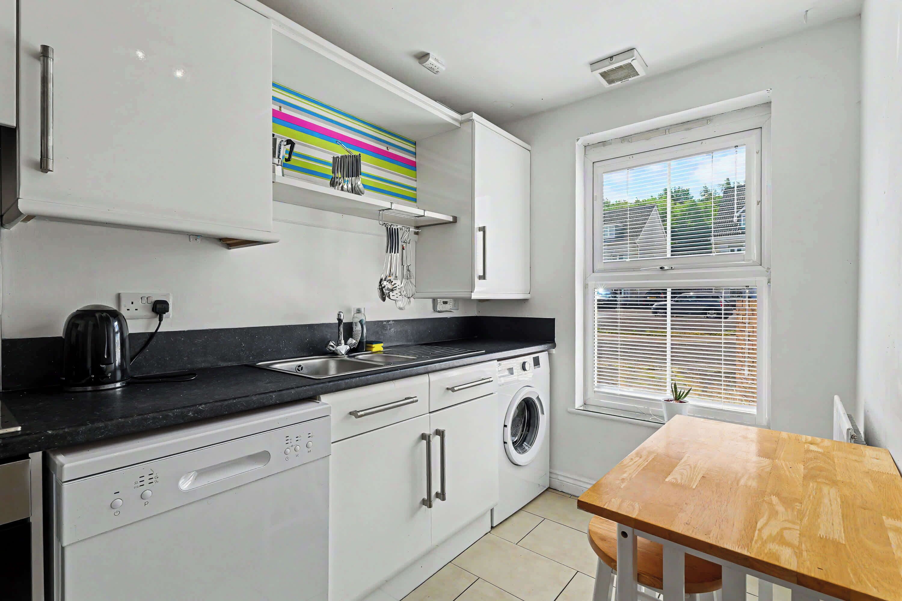 A modern kitchen with white cabinets, a black countertop, a washing machine, and a window with blinds.