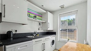 Een moderne keuken met witte kastjes, een zwart aanrecht, een wasmachine en een raam met zonwering.