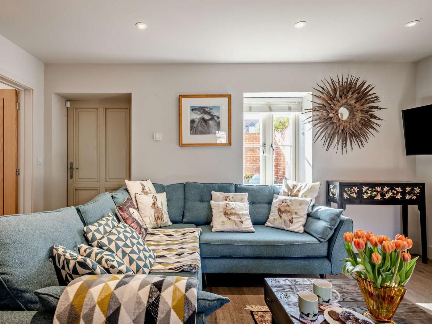 A living room with a sofa, patterned cushions, a coffee table with mugs, a vase of flowers, and a wall-mounted mirror.