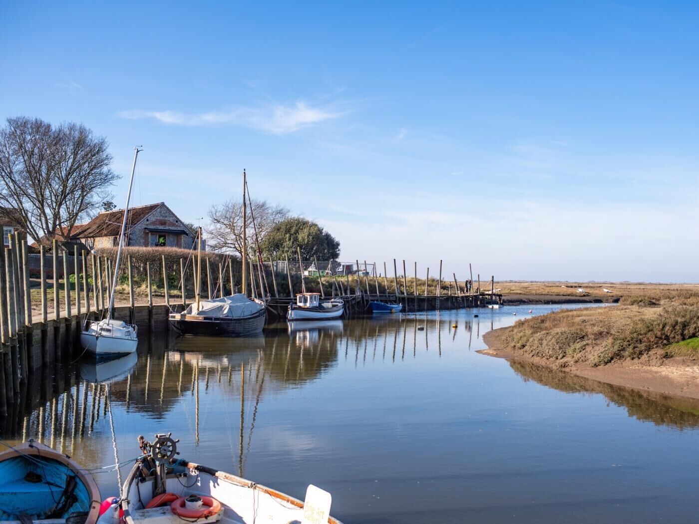 A calm waterway with boats docked, a wooden pier, and a house in the background.