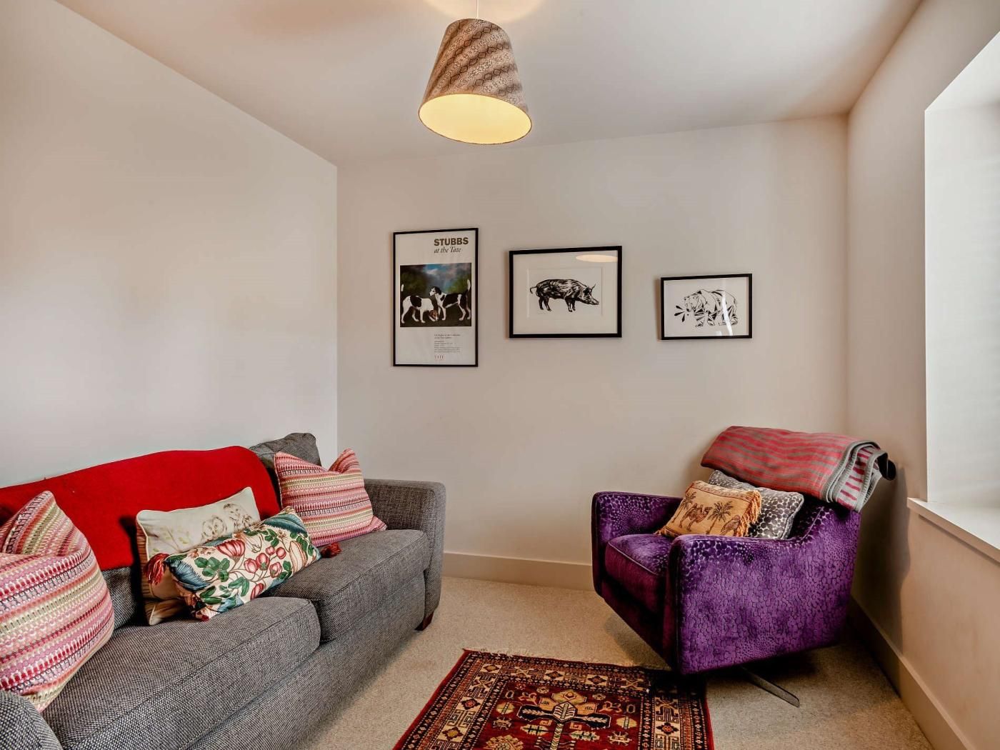 A living room with a grey sofa, a red throw blanket, a purple armchair, and a patterned rug. There are three framed pictures on the wall.