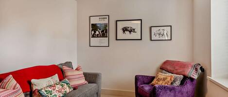 A living room with a grey sofa, a red throw blanket, a purple armchair, and a patterned rug. There are three framed pictures on the wall.