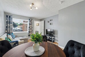Et spiseområde med et rundt bord, en vase med grønne planter, en sofa og et fjernsyn i baggrunden.