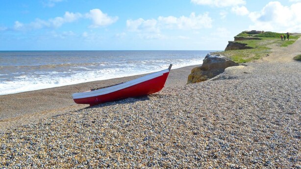 A pebbled beach with a red boat, a cliff with people, and the ocean. - 2 Bed in Holt (oc-ncc25) (Kelling)