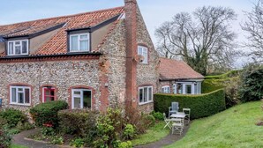 A stone house with a sloped roof, a red door, and a small garden with a table and chairs. - 2 Bed in Holt (oc-ncc25) (Kelling)