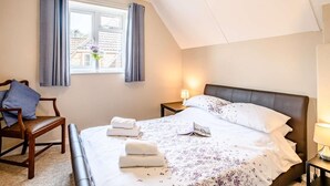 Ein Schlafzimmer mit Bett, Sessel, Nachttisch, Lampe und einem Fenster mit Vorhängen.
