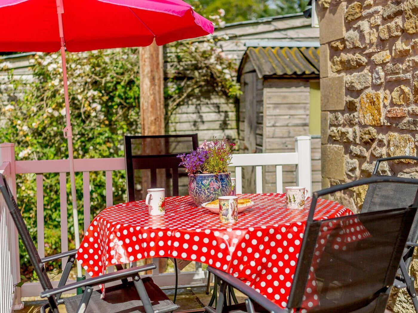 A patio with a red polka dot tablecloth, a vase with flowers, and a red umbrella.