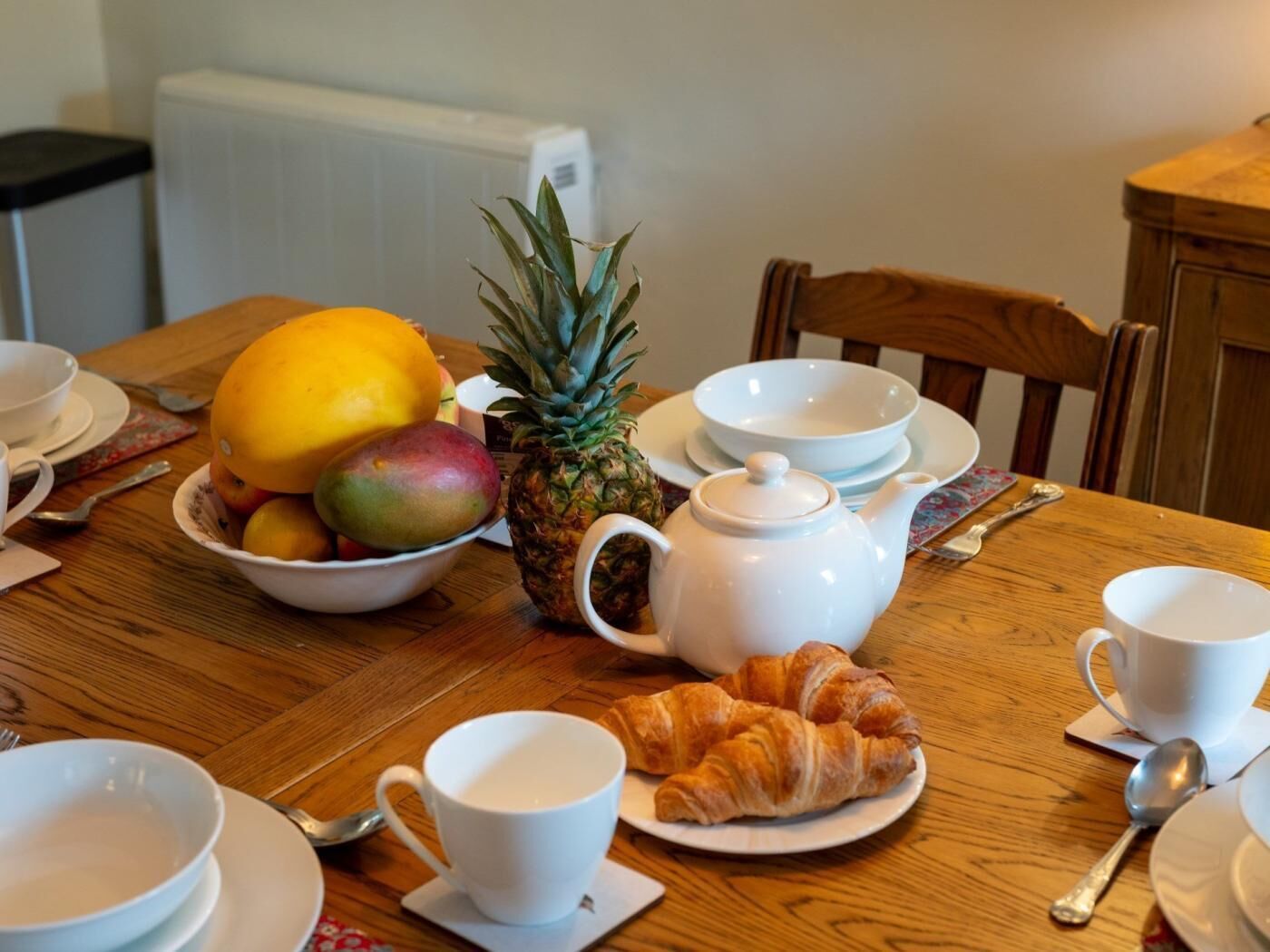 Une table en bois dressée pour le petit-déjeuner, avec un panier de fruits, un ananas, une théière et des croissants.