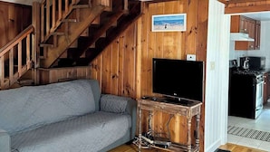 Un intérieur en bois avec un escalier, un canapé gris, une console en bois sur laquelle se trouve un téléviseur, et un coin cuisine.