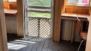 A wooden cabin with a porch, a door leading outside, and a window with a view of a yard and trees.