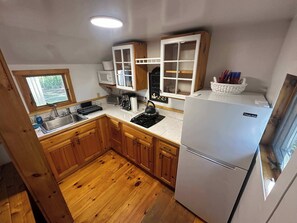 A compact kitchen with wooden cabinets, a stainless steel sink, a white refrigerator, and a window overlooking greenery. - Cozy 1 Bedroom Cabin on Campground/Retreat (Yarmouth)