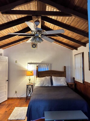 Un dormitorio con techo de madera, una cama con ropa blanca, una mesita de noche, una lámpara y una ventana con persianas.