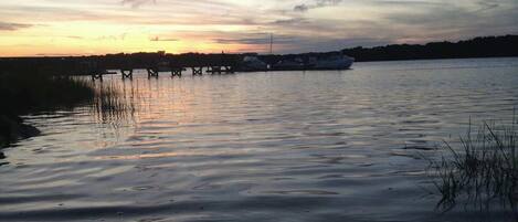 A calm waterfront with boats docked, a pier extending into the water, and a sunset in the background.