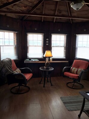 A cozy room with wooden walls, a wooden ceiling, and a window with blinds. There are two wicker chairs with cushions, a small round table with a lamp, and a wooden floor. - Charming 1 Bedroom Cottage on the Water! (South Dennis)