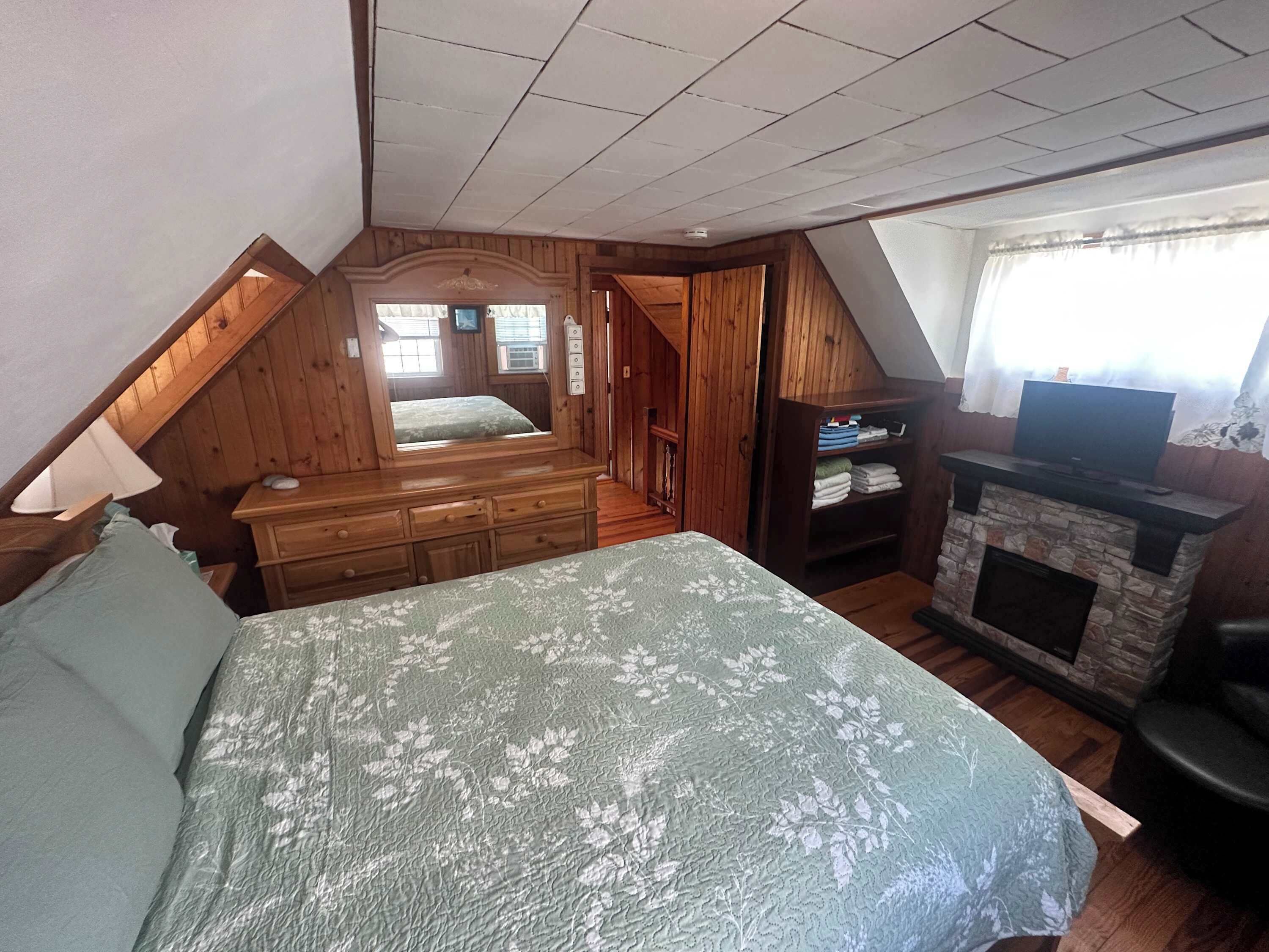 A bedroom with a sloped ceiling, a bed with a floral bedspread, a dresser, a vanity, a television, and a fireplace.
