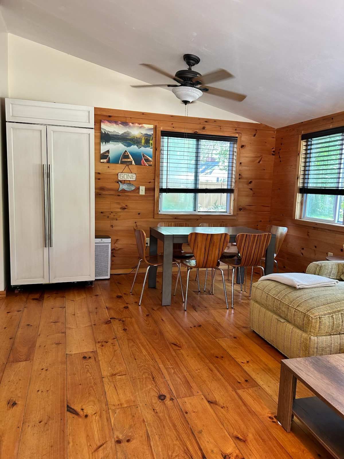 A room with wooden walls, a ceiling fan, a dining table with chairs, a bed, and a window with blinds.