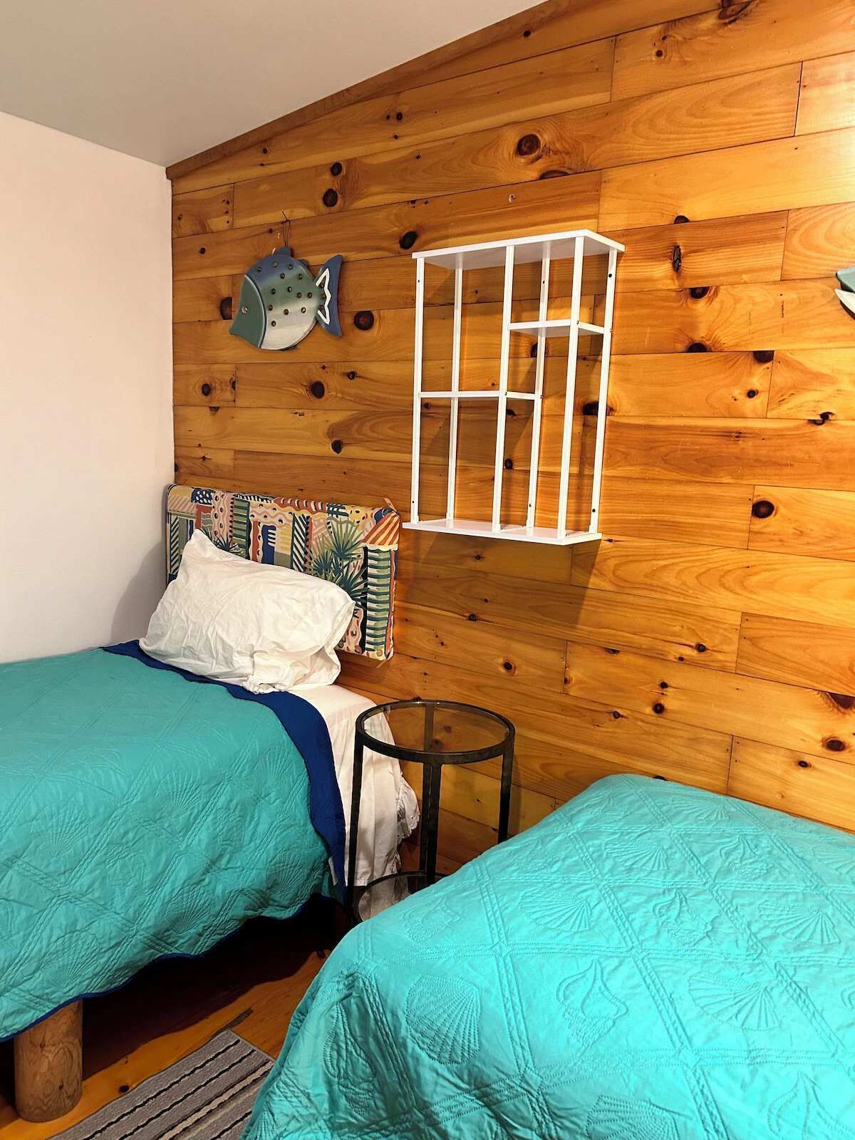 A room with wooden walls, two beds with teal bedding, a small round table, and a white hanging chair.
