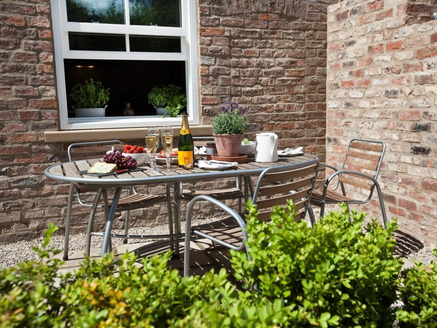 Une terrasse extérieure avec une table dressée pour deux personnes, une bouteille de champagne, un saladier de fraises et une plante en pot.