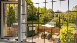 Blick durch ein Fenster mit Gittermuster, eine Terrassenfläche mit Gartenmöbeln und ein Garten mit verschiedenen Pflanzen und einem Holzzaun.