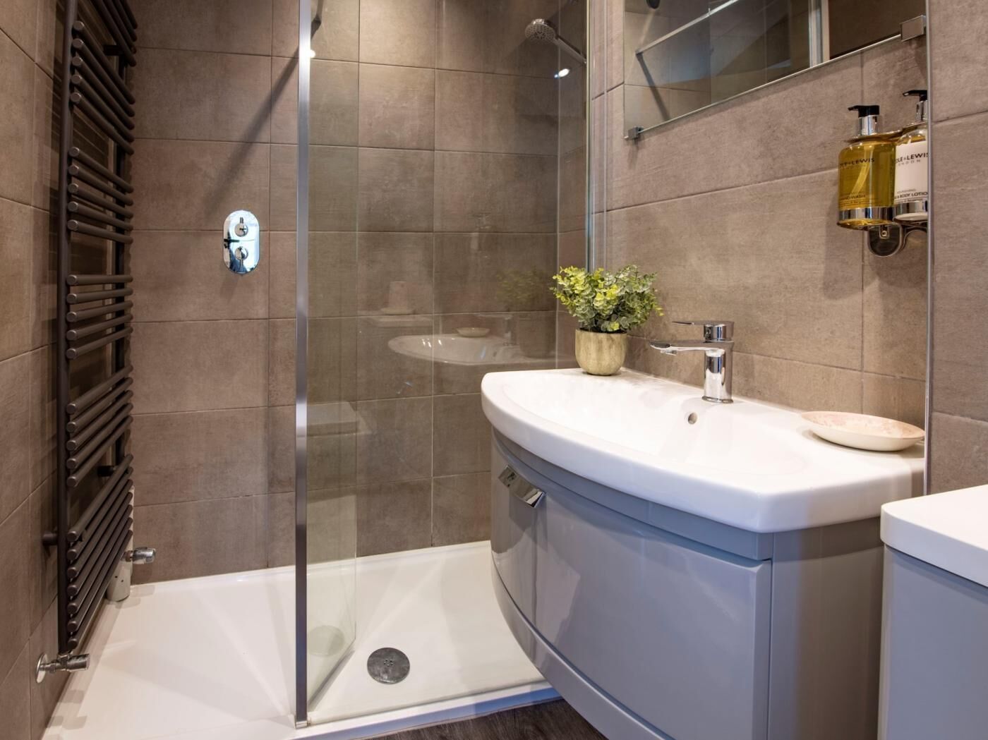 Un baño moderno con cabina de ducha de vidrio, un lavamanos blanco con una planta y artículos de aseo en la pared.