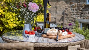 Ein runder Tisch aus Korbgeflecht mit kariertem Tuch, eine Weinflasche, ein Glas, ein Brotlaib, ein Messer, eine Tomatenschale und eine Vase mit Blumen.