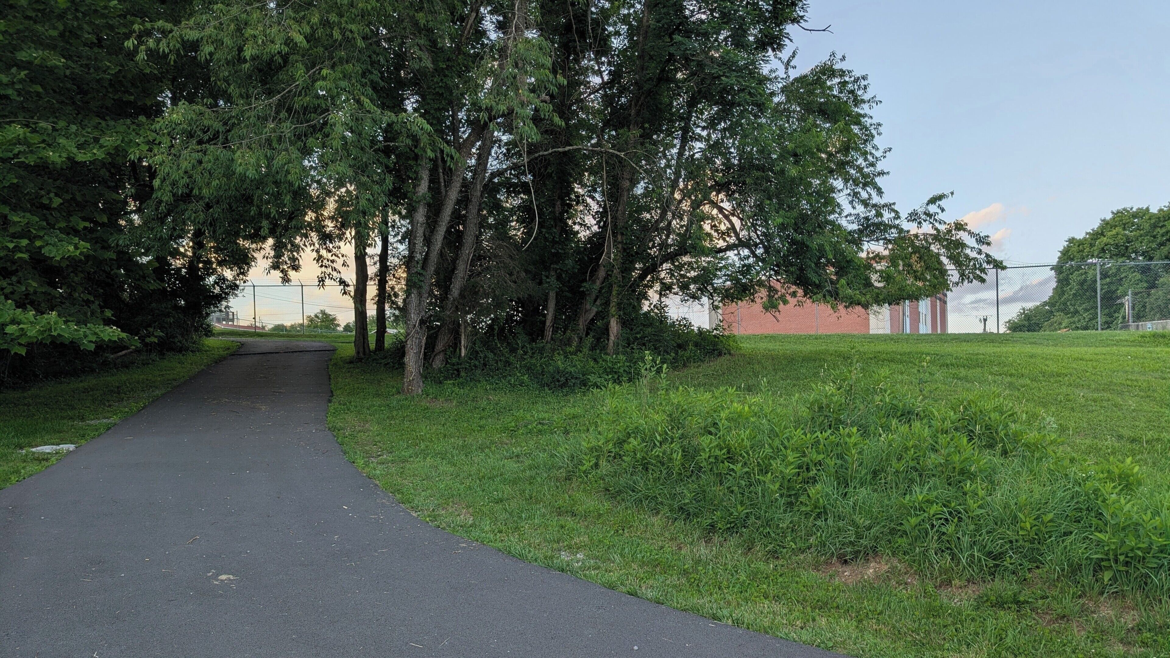 Asfaltoitu kävelytie kulkee nurmikon läpi, ja toisella puolella on puita ja taustalla rakennus.