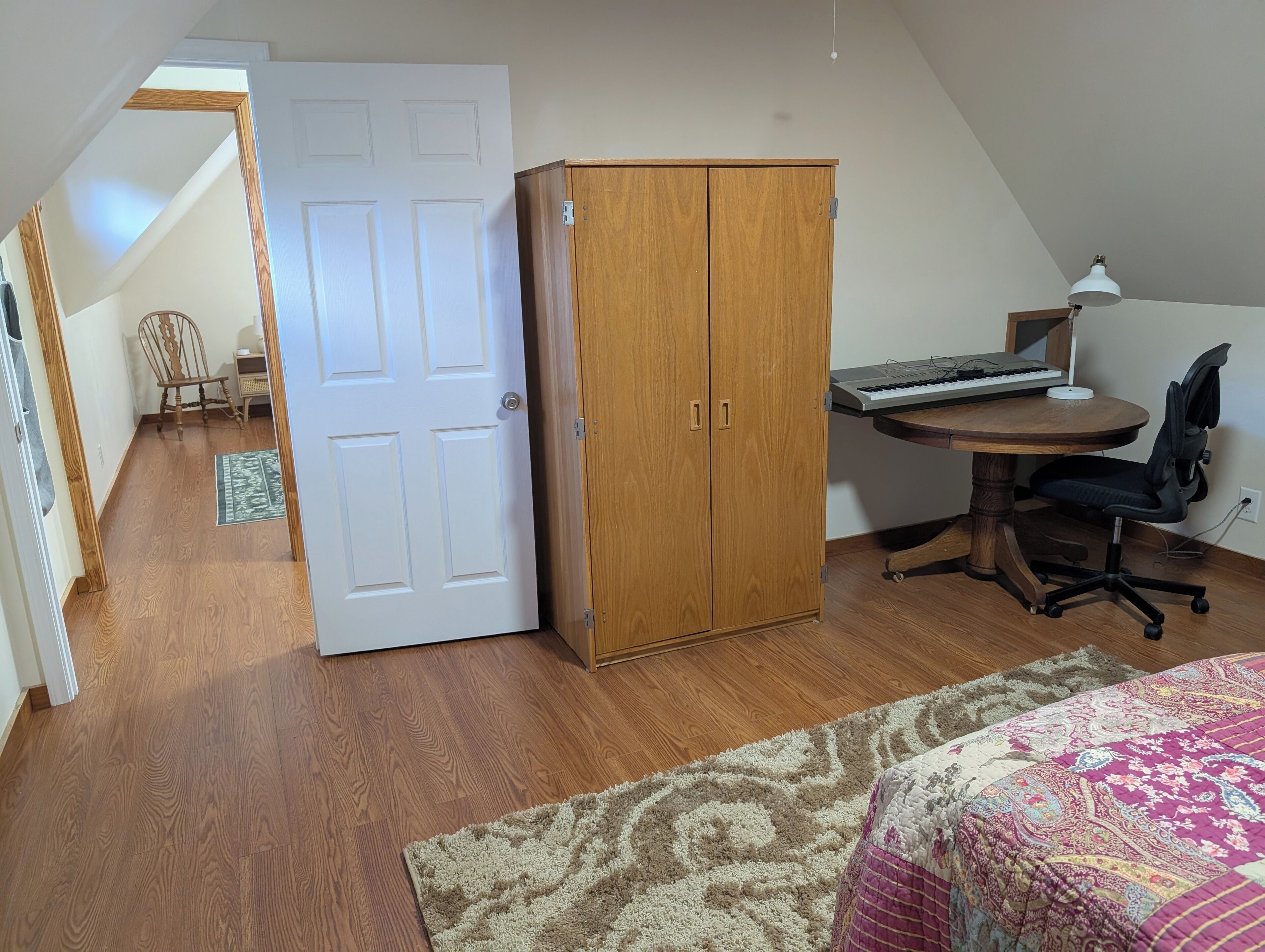 A room with a wooden wardrobe, a desk with a keyboard, a chair, and a bed with a patterned bedspread.