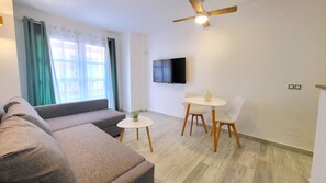 A modern living room with a sofa, a small table with chairs, a ceiling fan, and a flat-screen TV mounted on the wall. - Sol Suites 1ºPlus Apartment (Fuengirola)