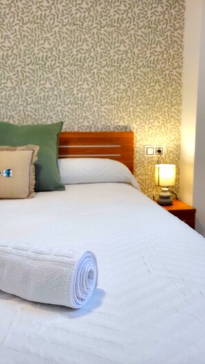 A neatly made bed with a rolled white towel, a wooden headboard, and a nightstand with a lamp. - Sol Suites Apartment 1ºPlus (Fuengirola)