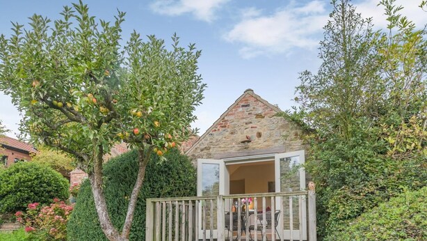 A small stone cottage with a wooden deck, surrounded by lush greenery and fruit trees. - 1 Bed in York (oc-g0032) (Crayke)