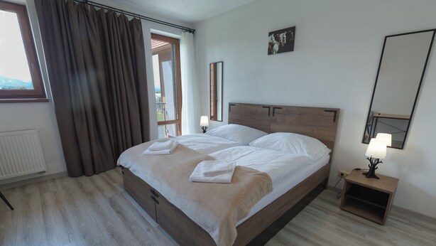 A hotel room with a bed, a bedside table with a lamp, a window with curtains, and a view of the outside. - Penzion Kamence (Trstene)