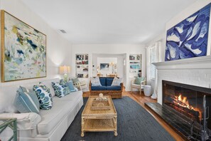 Un salon avec un canapé blanc, des coussins bleus, une table basse en osier, une cheminée et une bibliothèque.